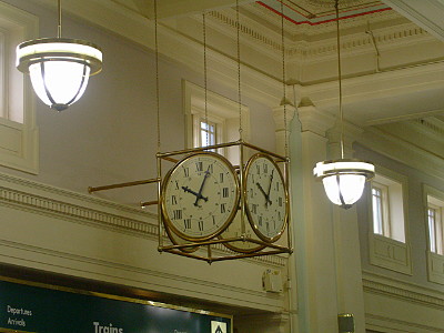 [An brass cubical frame has a clock circled in brass on each of its four sides with roman numerals on each clock. This structure hangs from the two story ceiling. The upper store has windows.]